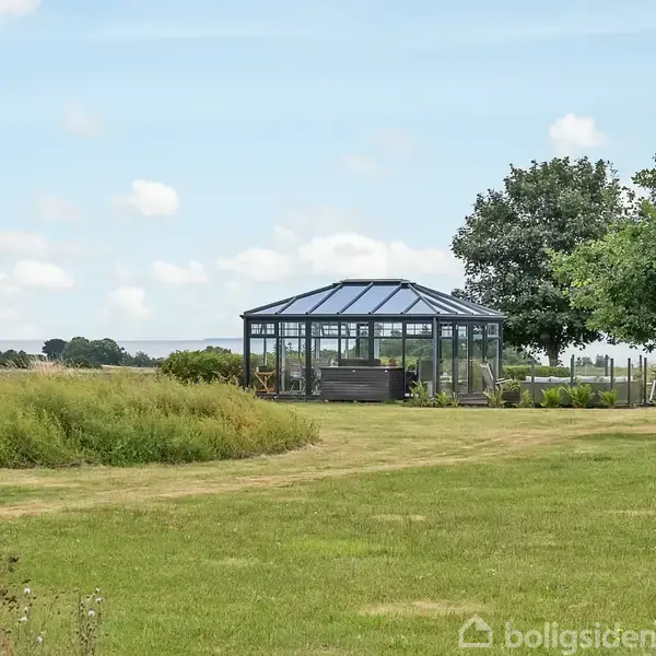Et drivhus står i en stor, åben græsset have ved siden af en træplantning. Skyer svæver i en klar, blå himmel over det landlige landskab.