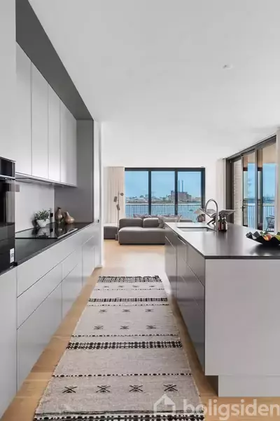 Et moderne køkken med hvide skabe og en sort bordplade har en lang løber på gulvet. I baggrunden ses en stue med en sofa og store vinduer med udsigt til vandet.