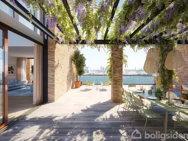 Terrassen er dækket med et pergolatak fyldt med grønne planter og blomster. Et langt spisebord står til højre, mens der i baggrunden ses en udsigt over vandet.