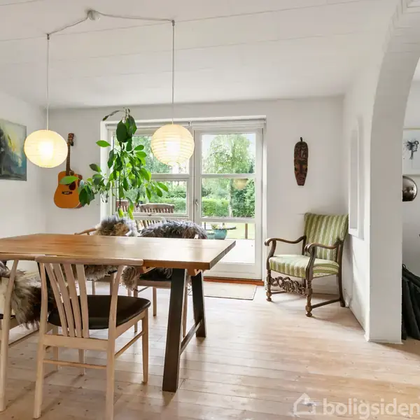 En spisestue med et træbord og stole, der er dækket af fåreskind, står centralt. To lamper hænger over bordet. En guitar hænger på væggen. Store vinduer viser en have.