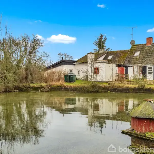 Et lille rødt andehus står stille på en grøn platform i en dam. I baggrunden ses et gammelt, hvidt hus med mos på taget omgivet af træer og klar blå himmel.