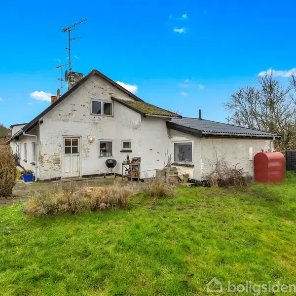 Et hus med hvid facade står i en baghave med grønt græs. Der er et skur til højre, buske til venstre, og klare blå himmel i baggrunden.