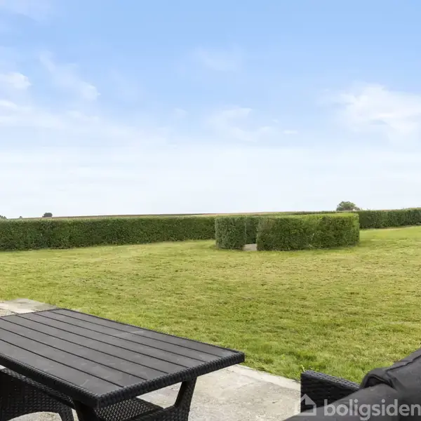 En sort bænk står på en terrasse foran en velplejet græsplæne omkranset af lave hække under en blå himmel.