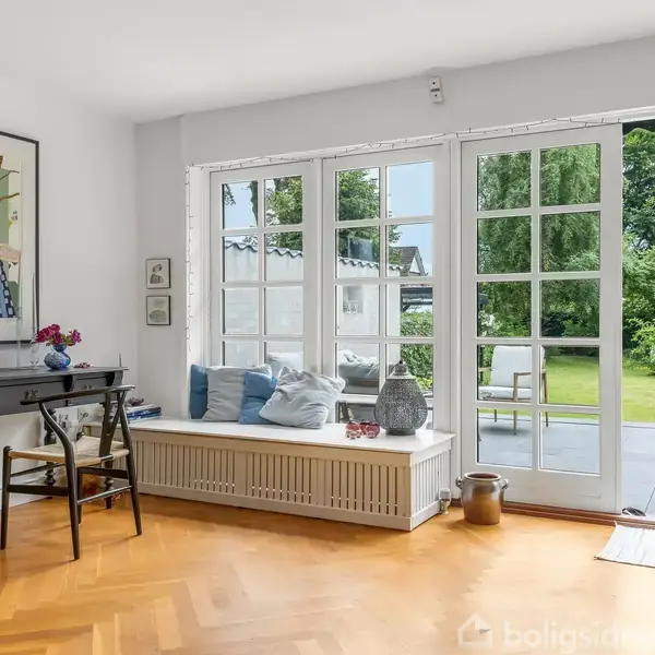 Et elegant rum med et sort skrivebord og stol ved siden af en stor vinduesfront. Vinduerne åbner ud mod en terrasse med udsigt til en grøn have.