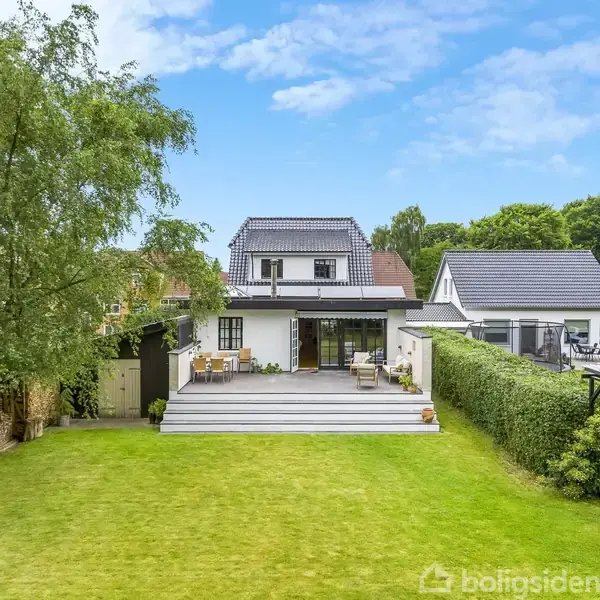 Et hvidt to-etagers hus står i en velplejet have med grøn græsplæne. En træterrasse støder op til huset, omgivet af buske og store træer under en blå himmel.