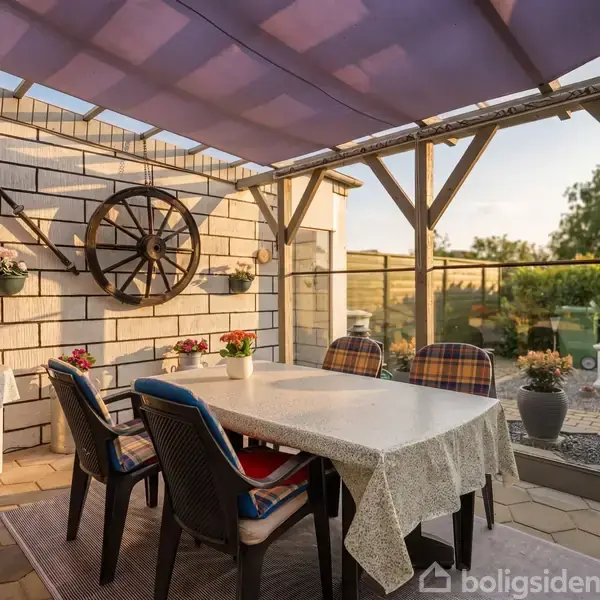 En overdækket terrasse med et bord dækket af en dug omgivet af stole. En vognhjul og planter dekorerer væggen. Haven ses i baggrunden med græs og buske.