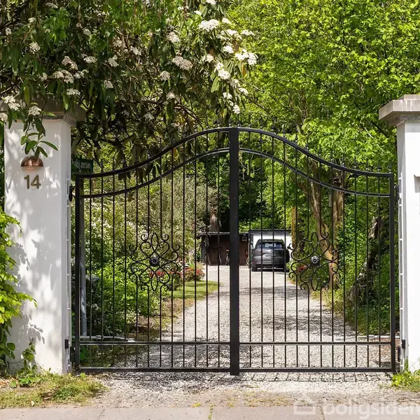 Sort smedejernslåge står åben og afslører en grusvej med en bil i det fjerne. Omgivet af frodig grøn vegetation og hvidmalede søjler, nummereret 14 og med en postkasse.