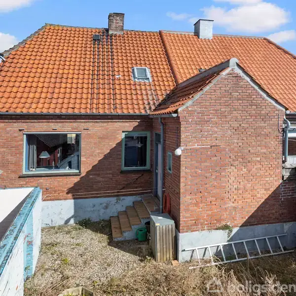 Rødt murstenshus med orange tegltak, to vinduer og skorsten midt i billedet. Omgivet af et lille, indhegnet udendørsområde med trapper og en stige liggende mod muren.