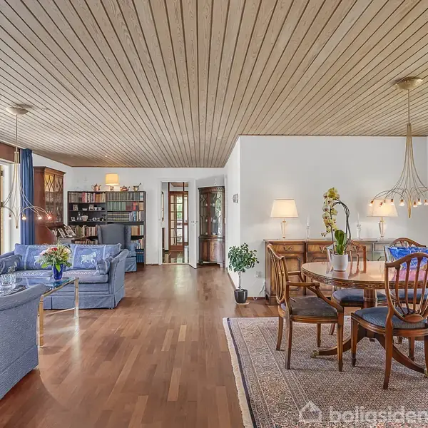Stue med blå sofaer og et sofabord; spisestue med træbord og stole. Trægulv og -loft, reoler langs væggen. Store vinduer giver naturligt lys.