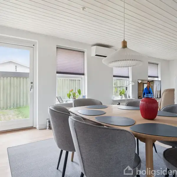 Rundt spisebord med otte grå stole står i en stue, dækket med dækkeservietter og en rød vase. Store vinduer lukker lys ind, og en person ses ved bagvinduet.