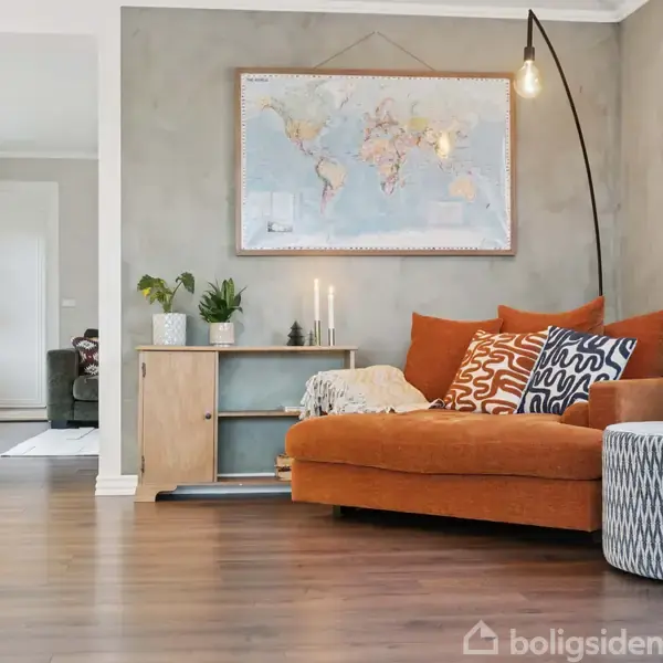 Orange sofa med mønstrede puder står i en stue, oplyst af en gulvlampe. Verdenskort hænger på væggen over sofaen. Trægulv og planter i potter fuldender rummet.