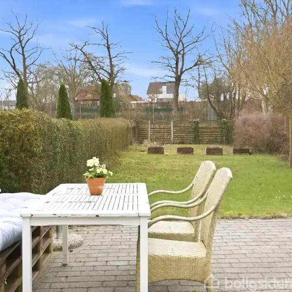 Udendørsmøbler står på en flisebelagt terrasse; en hvidmalet bord og kurvestole ved siden af en sofa lavet af træpaller. Haven er omgivet af hække og træer under en blå himmel.