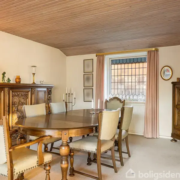 Et rundt træbord omgivet af seks polstrede stole står i en spisestue. I baggrunden ses et træskab, en lampe, kunstgenstande og et vindue med glasrude.