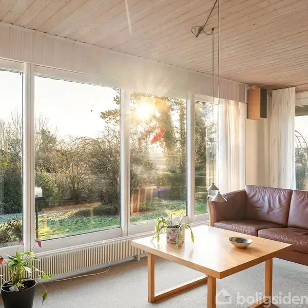 En stue med en brun lædersofa ved siden af et træbord. Store vinduer lader sollys skinne ind og viser en udsigt over en have med træer.