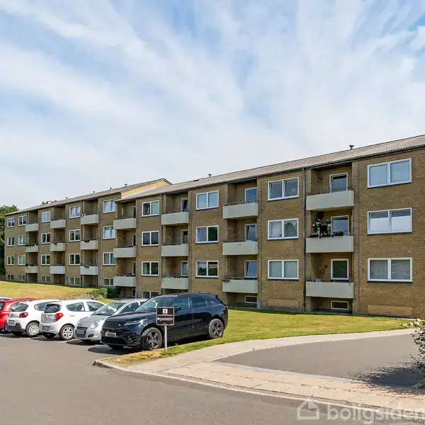 Et lejlighedskompleks i flere etager står stille midt i et grønt område. Biler er parkeret foran bygningen på en asfalteret parkeringsplads. Himlen er blå og delvist skyet.