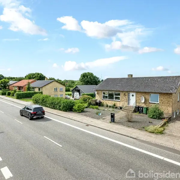 Et parcelhus med gul murstensfacade og sort tag står roligt ved en landevej, hvor biler kører forbi. Omkring huset er der lavt beplantet have og grønne marker i baggrunden.