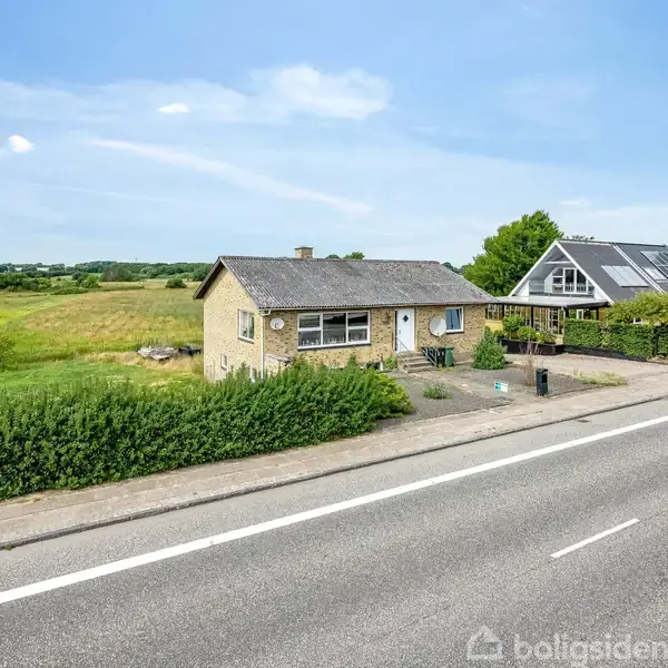 Et gult murstenshus med gråt tag står ved siden af en bred vej. Omkringliggende grønne marker strækker sig i det åbne landskab under en blå himmel.