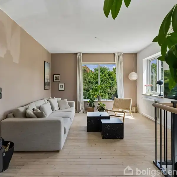 En grå sofa står langs en væg ved siden af en tændt pejs i en moderne stue. Store vinduer giver udsigt til grønne omgivelser. En plante står nær en trappe.