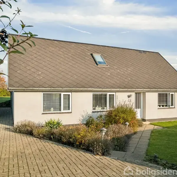 Et hvidt hus med skråt tag står stille, omgivet af en have med buske. En bred flisebelagt indkørsel fører op til huset.