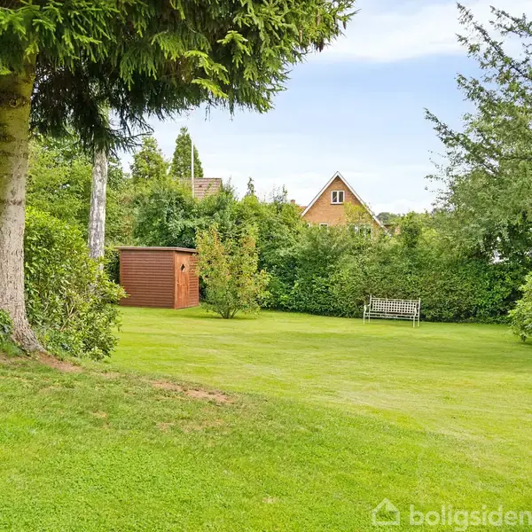 Græsplæne med en brun skur, omgivet af træer og buske. En hvid bænk står ved en hæk. I baggrunden ses taget af et hus.