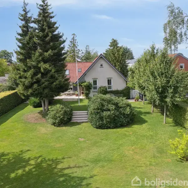 Et hvidt hus med rødt tag står i en velplejet have med store træer og buske. Stentrin forbinder terrassen til den grønne plæne.