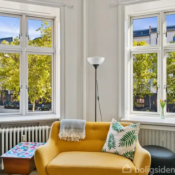 Gul sofa står foran to lyse vinduer med grønne træer udenfor. En standerlampe står ved siden af sofaen, med puder og et tæppe. Møblementet skaber en hyggelig stueatmosfære.