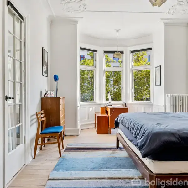 Soveværelse med dobbeltseng til højre, skrivebord foran vinduer i karnap, og skab med glasdøre til venstre. Rum med lyse vægge og træmøbler, grønt udenfor vinduerne.