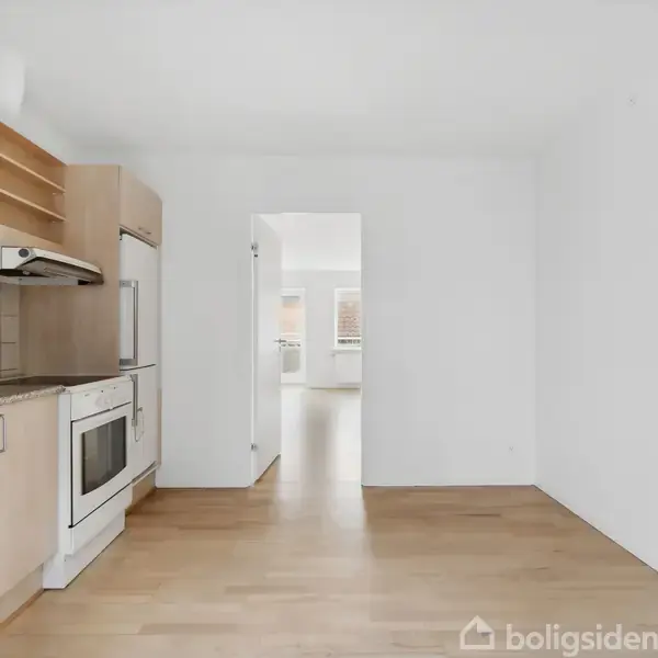 Køkken med træelementer og hvidevarer, tomt og lyst med åbning til tilstødende rum i et minimalistisk hjemmemiljø.