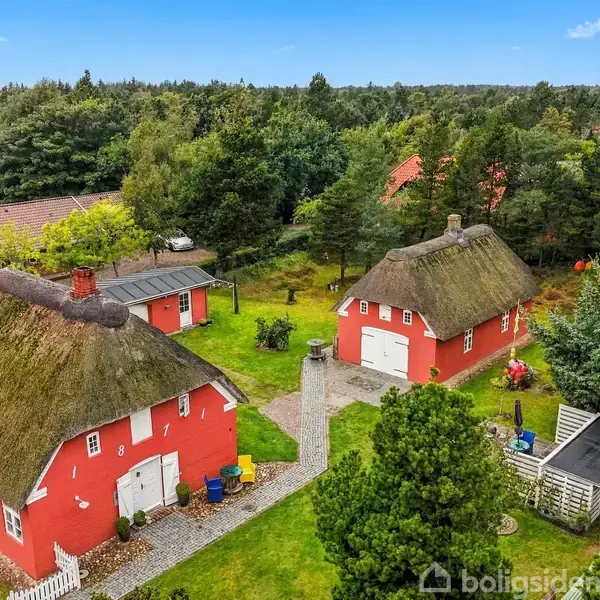 To røde huse med stråtage står tæt på hinanden. De er omgivet af en frodig have med grønne træer og buske, og der er en lille sti imellem dem.