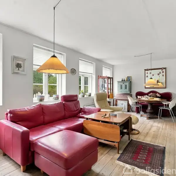 Rødt sofaarrangement placeret i en lys stue med trægulve. Sofaen vender mod et sofabord og spisebordområde. Fire vinduer lader naturligt lys ind i rummet.