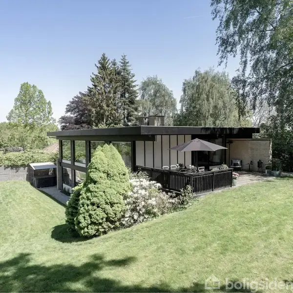 Et moderne hus med fladt tag ligger i en frodig have. Huset har en stor terrasse med parasol. Træer og buske omringer ejendommen, skaber en fredfyldt atmosfære.