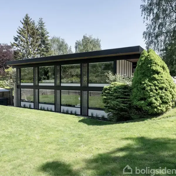 En moderne, kubisk bygning står stille i en velplejet have med græsplæne og buske, omgivet af høje træer, hvilket giver et naturligt og fredfyldt miljø.