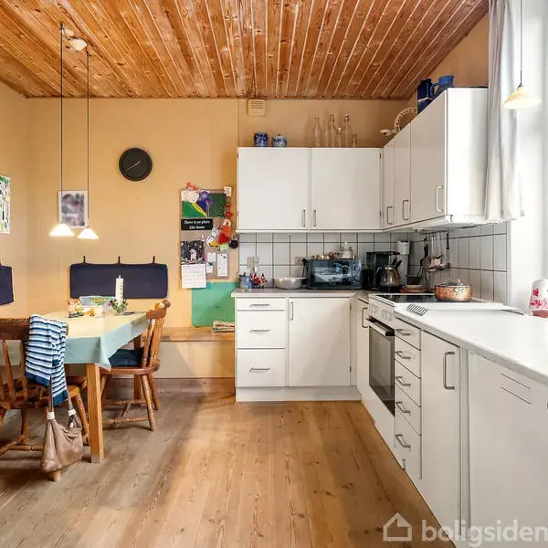 Køkken med træbord og stole, dækket med dug, i en lys spisestue. Hvide skabe og køkkenudstyr fylder det højre område. Vinduer giver naturligt lys fra højre.
