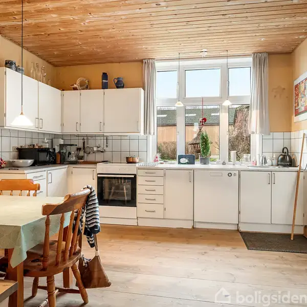 Et køkken med hvide skabe og ovn, udsmykket med børnetegninger. Et spisebord med træstole står i forgrunden, mens et stort vindue i baggrunden giver naturligt lys.
