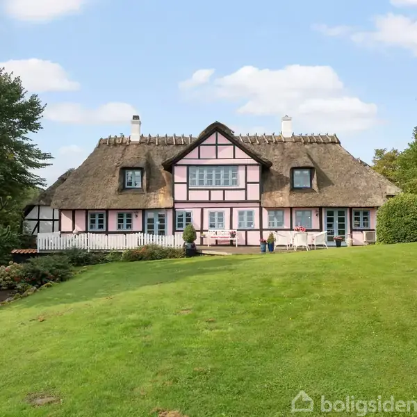 Hus med stråtag står på en grøn plæne. Det har hvidt og lyserødt bindingsværk. Omkringliggende træer og buske omkranser bygningen i en have.