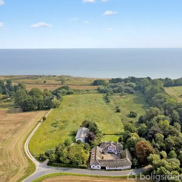 En stor gård omgivet af marker og skov ligger i et åbent landskab nær kysten, med havet i horisonten under en klar blå himmel.