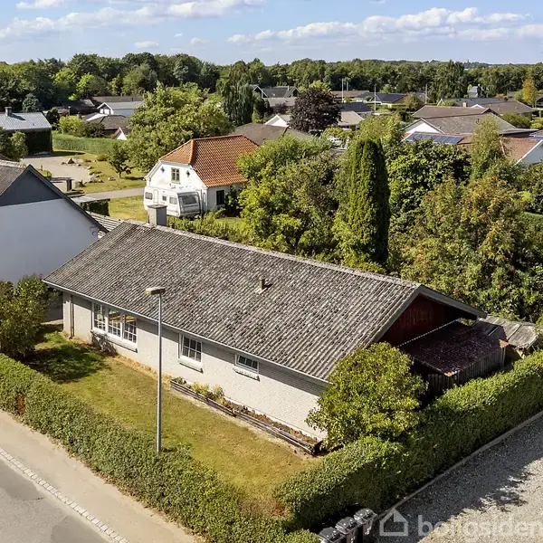 En enkelt-etagers hvid bygning med gråt tag er omgivet af en hæk og ligger ved siden af andre huse i et grønt boligområde med træer og buske.
