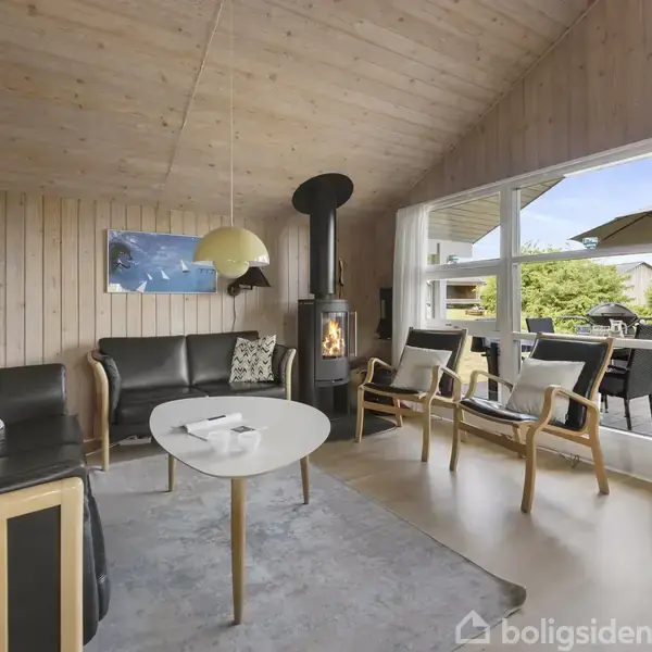 Stue med sorte lædersofaer og stole omkring et hvidt bord; pejs i hjørnet. Store vinduer åbner til en terrasse med møbler og udsigt til grønt område.