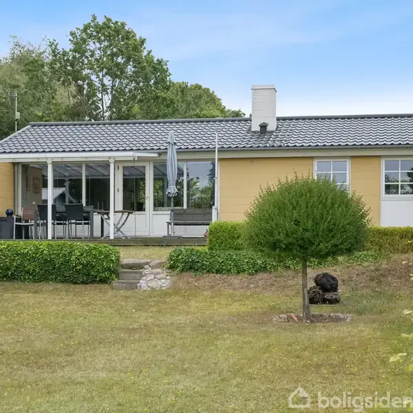 En gul murstensbungalow med sort tag står på en velplejet græsplæne med en terrasse og havelåge. Haven er omgivet af buske og træer i baggrunden.