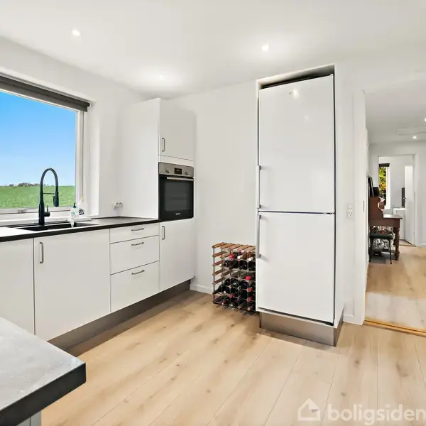 Et lyst, moderne køkken med hvide skabe og indbygget ovn står stille. En stor vindue giver udsigt til en grøn mark. Rummet strækker sig ud mod et spiseområde.