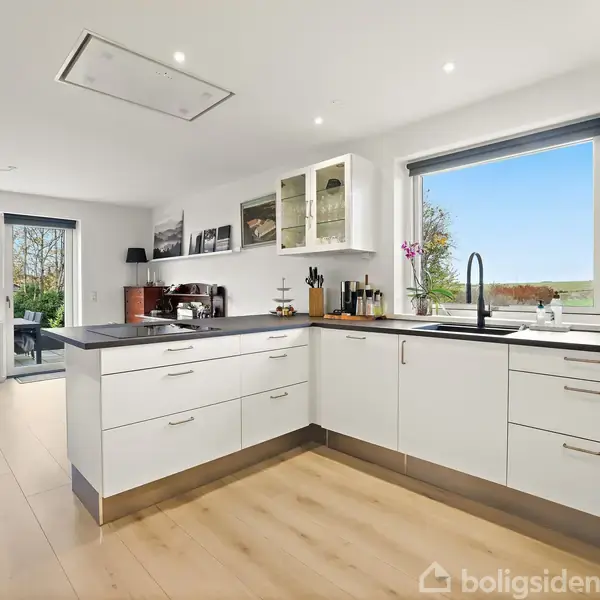 Et moderne, lyst køkken med hvide skabe og sorte bordplader. Store vinduer giver udsigt til grønt landskab. I baggrunden ses en spiseplads med træbord og udesigt.