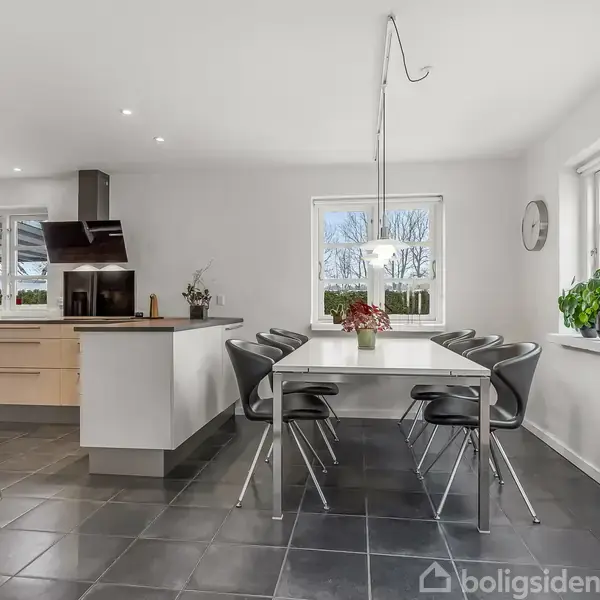 Et moderne køkken-alrum med sort flisegulv. Et hvidt spisebord med sorte stole står i rummet. Køkkenet har lyse træskabe og sorte apparater, mens store vinduer giver lysindfald.