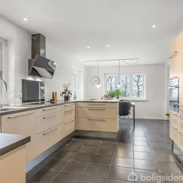 Køkken med lyse træskabe og mørkt flisegulv. En køkkenø er centralt placeret, omgivet af vinduer, der giver udsigt til træer udenfor. Møbleret med moderne apparater.