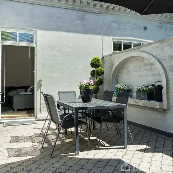 En terrasse med bord og fire stole er placeret på fliser. Bordet har en blomsterpotte. Døren til stuen står åben, og en mur omkranser området.