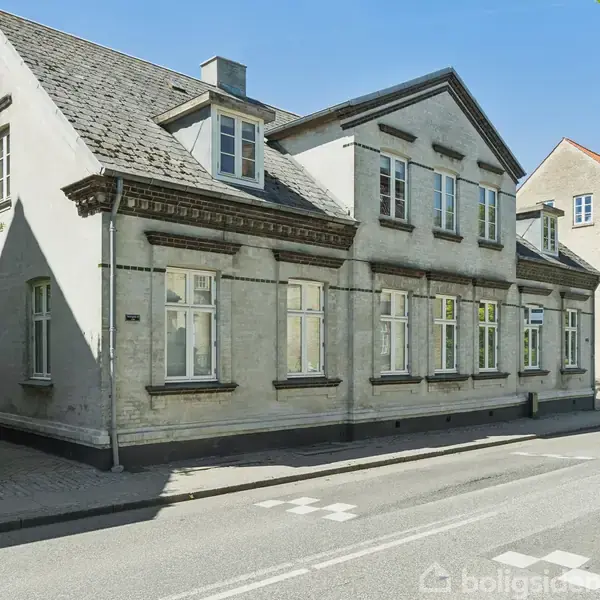 En hvid murstensbygning med skifertag står på en gadehjørne. Bygningen har flere store vinduer, og en skilt ses i venstre side. Gaden er stille og beliggende i et bymiljø.