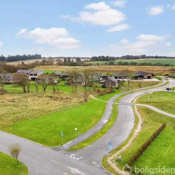 En vej krydser en grøn landskab med spredte huse i baggrunden under en blå himmel med hvide skyer. Ingen tekst er til stede.