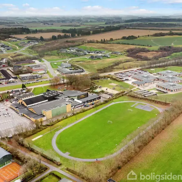 Fugleperspektiv af en skole med runde sportsbaner og flere bygninger omgivet af landbrugsjord og skove, i en rolig landområde.