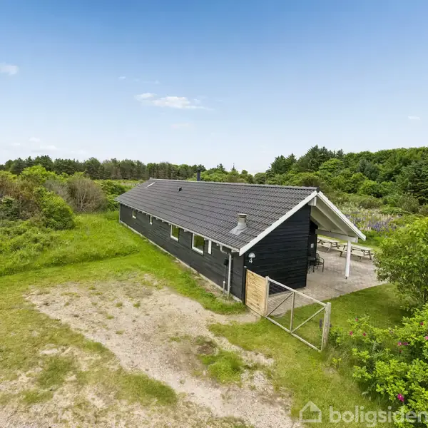 Et sort træhus ligger stille omgivet af et grønt, frodigt buskads under en klar blå himmel. Grusstier snor sig gennem landskabet omkring huset.