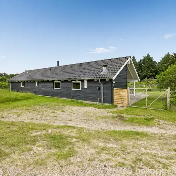 Sort træhus står stille i en grøn, skovklædt omgivelser. Huset har et tag med grå tegl og en åben indgang gennem en trælåge. Lys blå himmel over.