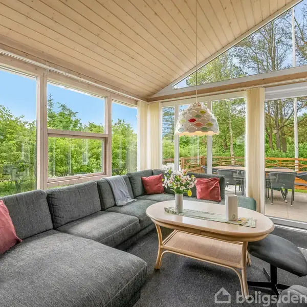 Grå hjørnesofa med røde puder står ved store vinduer og glasdøre. Et sofabord med vase og bøger står foran. Udenfor ses terrasse og frodig have.